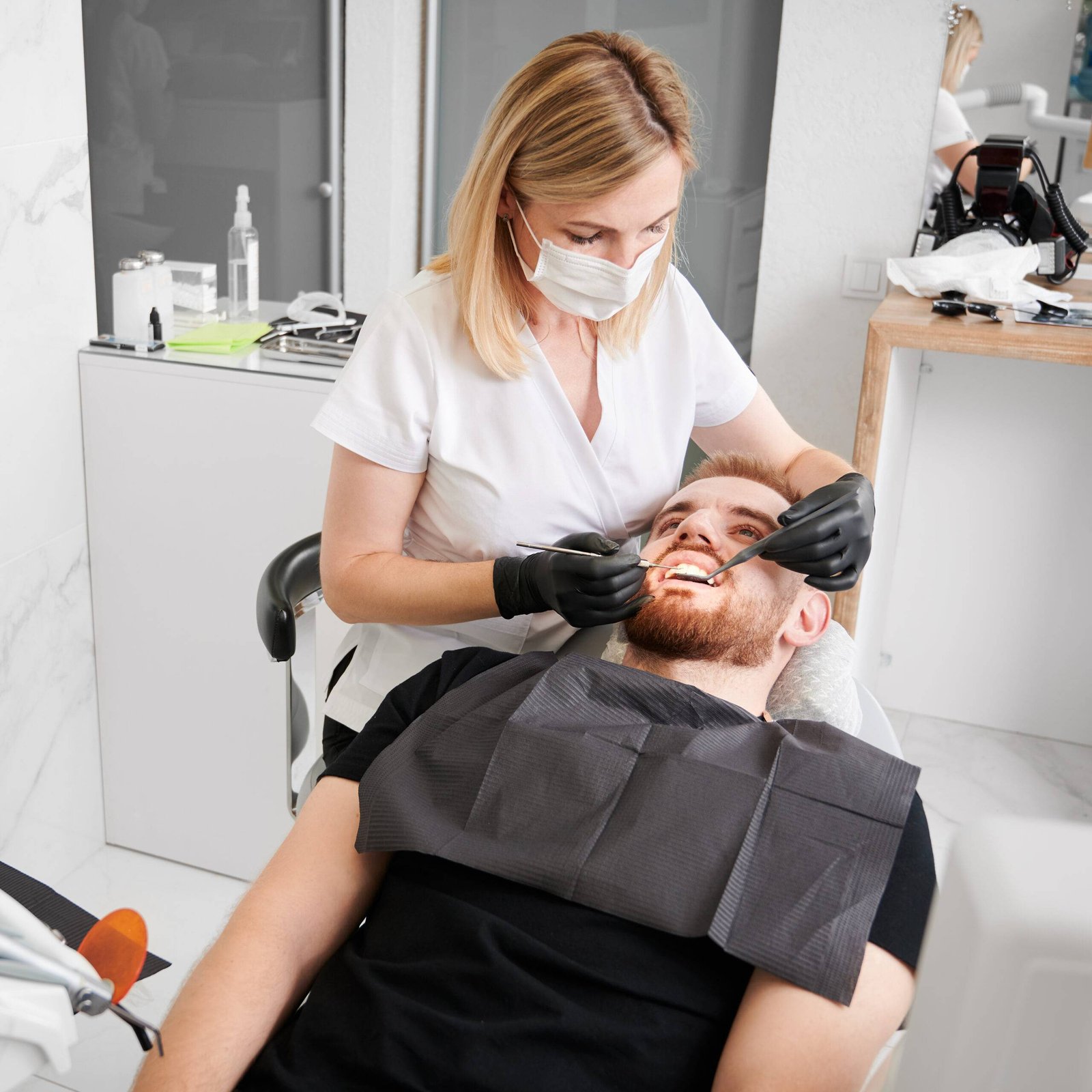 Overhead view on patient lying on dentist chair, wearing black bib, female dentist performing teeth check up with dental tools, wearing black gloves, face mask and uniform. Concept of dentistry
